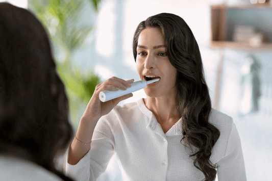 Woman using a sonic toothbrush while looking in the mirror, demonstrating effective teeth brushing technique.